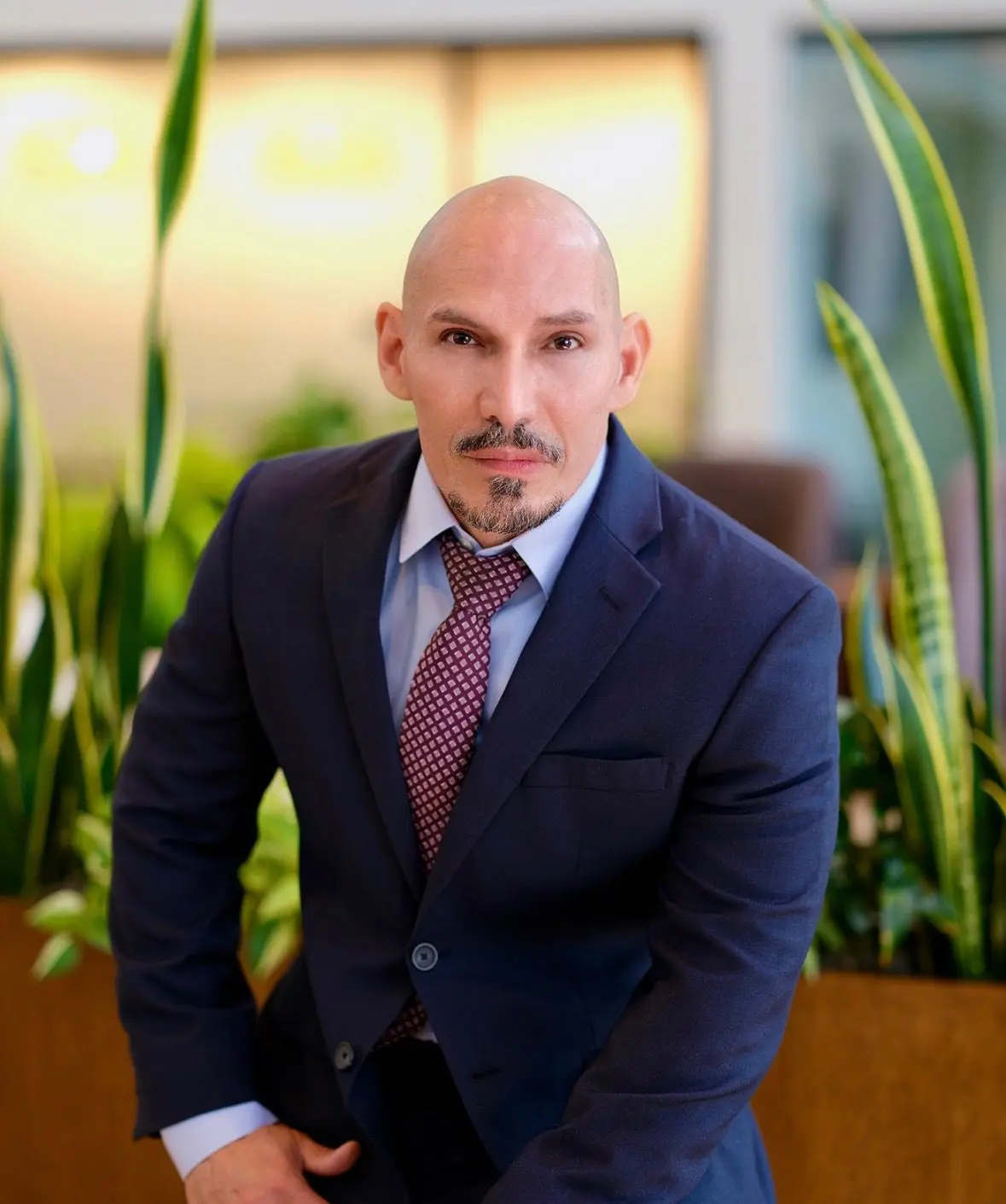 A confident man in a suit posing indoors with plants in the background.