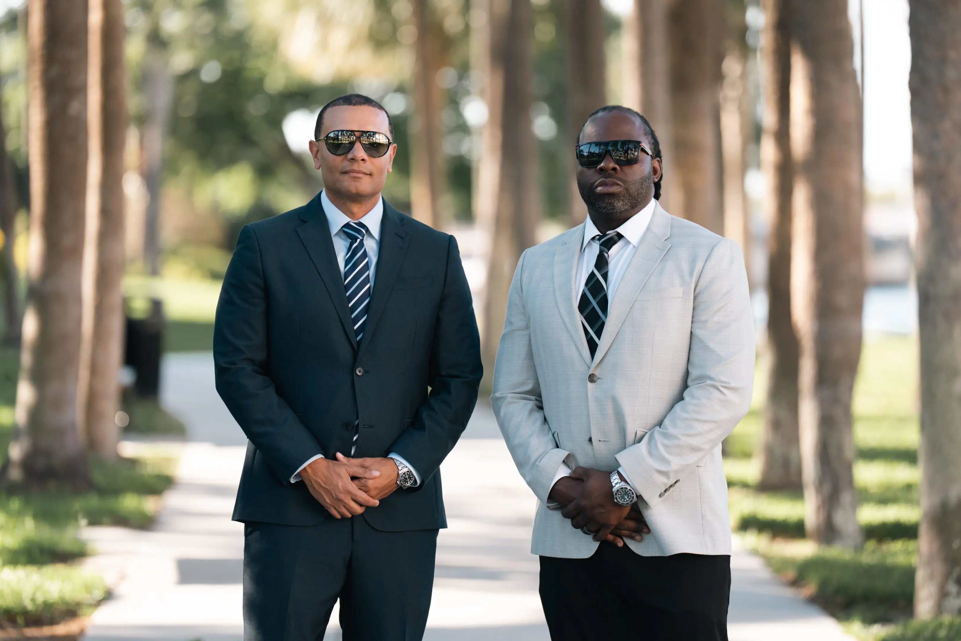 Two men in suits and sunglasses standing outside with trees in the background.