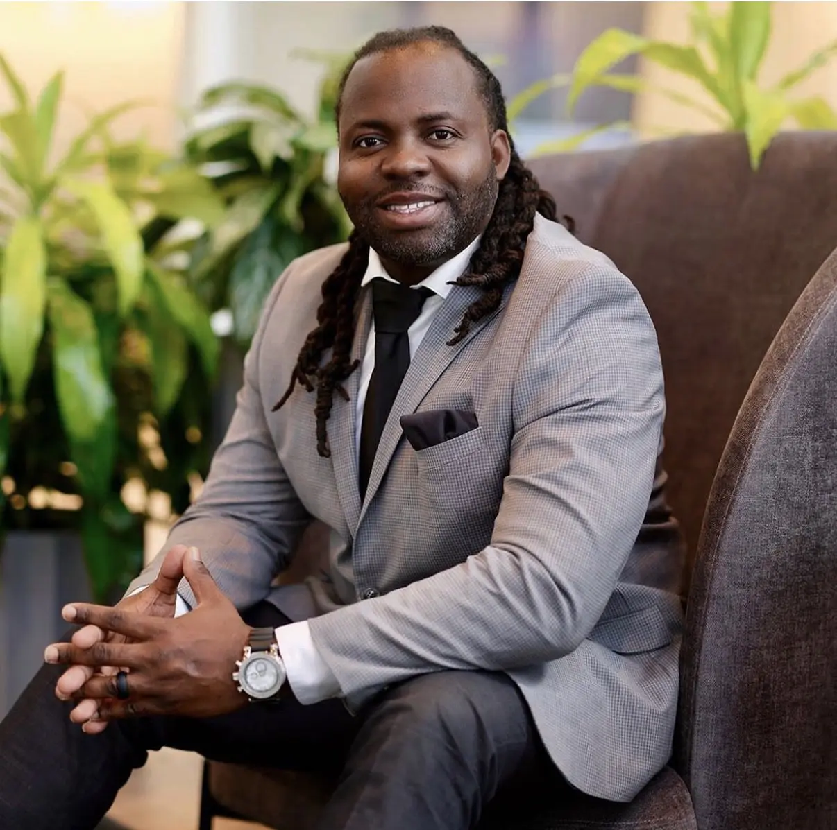 A man in a gray suit sitting on a chair, smiling confidently.