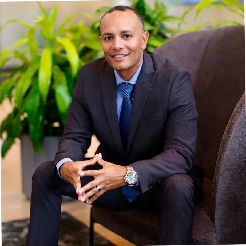 A man in a suit sitting on a sofa, smiling confidently.